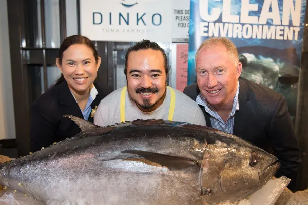 Three smiling people posing behind a very large southern bluefin tuna displayed on ice, with Dinko Tuna branding visible in the background.