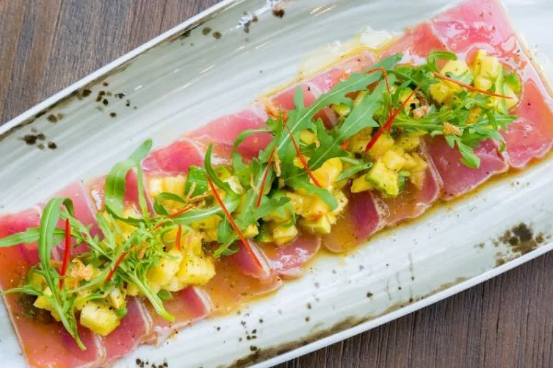 Sliced tuna carpaccio topped with diced pineapple salsa, rocket and a light dressing, arranged neatly on a long ceramic plate.