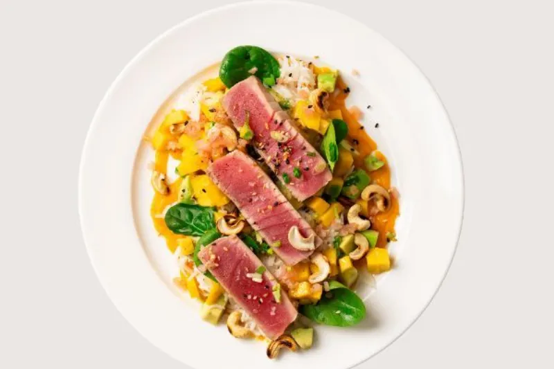 Seared tuna slices served over rice with mango, avocado, cashews and greens on a white plate.