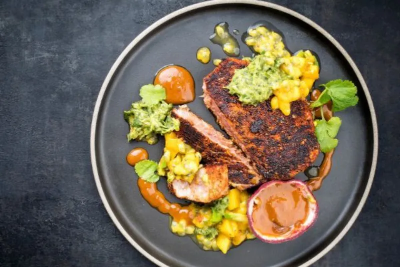 Cajun-crusted tuna steak plated with vibrant mango salsa, green herb sauce and passionfruit puree.