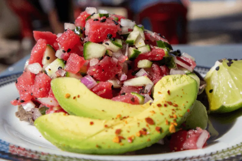 Tuna ceviche mixed with cucumber, onion and herbs, served with fresh avocado slices and lime on a plate.