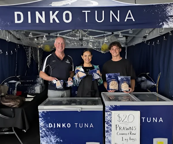 Three people standing behind a Dinko Tuna seafood stall at a market, holding packaged prawns and seafood products.