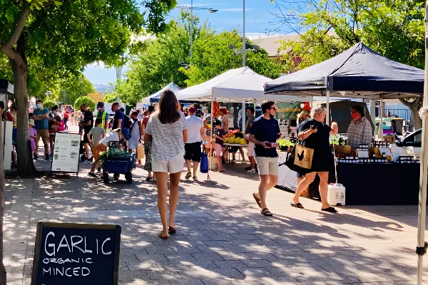 People walking and shopping at an outdoor farmers market with stalls selling fresh produce and food.