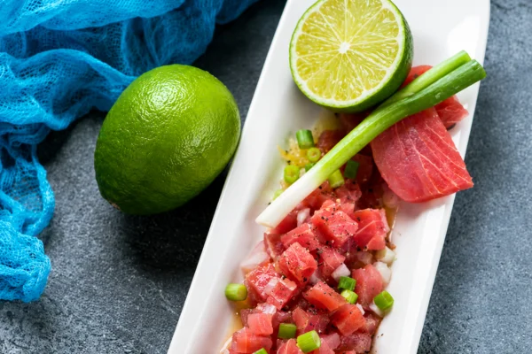 Rectangular white plate with tuna tartare, chopped green onions, and a lime half, garnished with a green onion stalk on a dark surface with a blue net nearby.