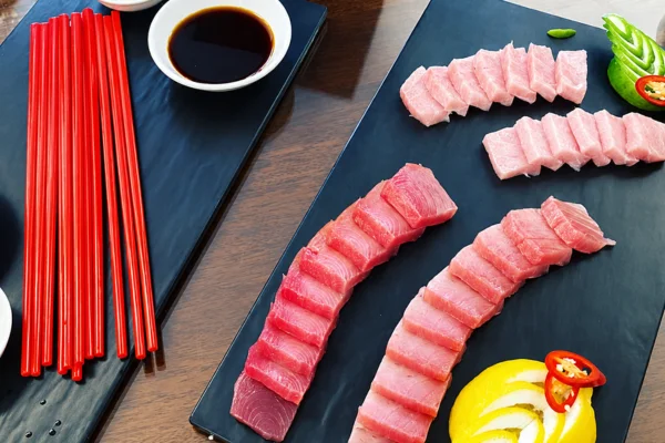 Assorted sashimi slices arranged on black boards with soy sauce, lemon wedges, and red chili peppers; bright red chopsticks rest nearby.