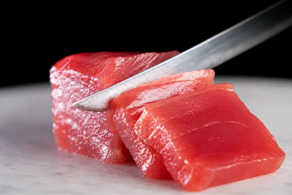 Two bright red tuna steaks on a cutting board as a knife slices through one piece, ready to cook.