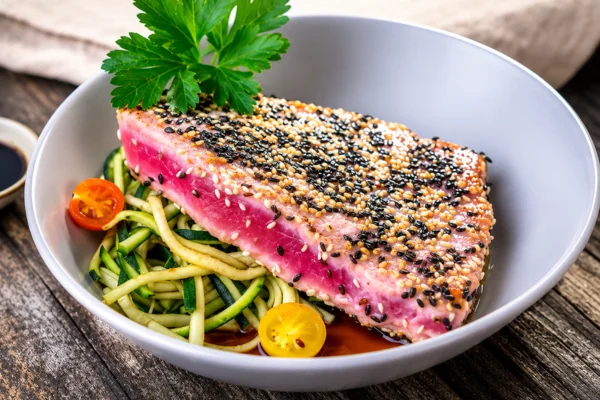 Sesame-crusted tuna steak with pink center on zucchini noodles and cherry tomatoes, drizzled with soy sauce and parsley.